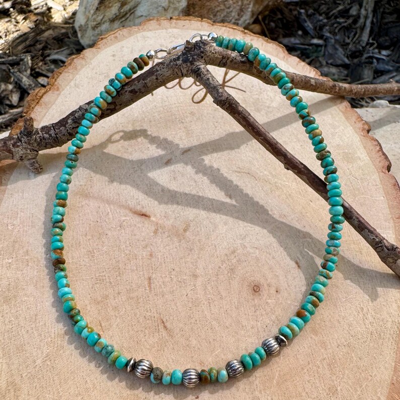 May include: A turquoise beaded necklace with silver accents. The necklace features small, round turquoise beads interspersed with silver-colored, textured beads. The necklace is displayed on a wooden surface, showcasing its natural and earthy aesthetic.