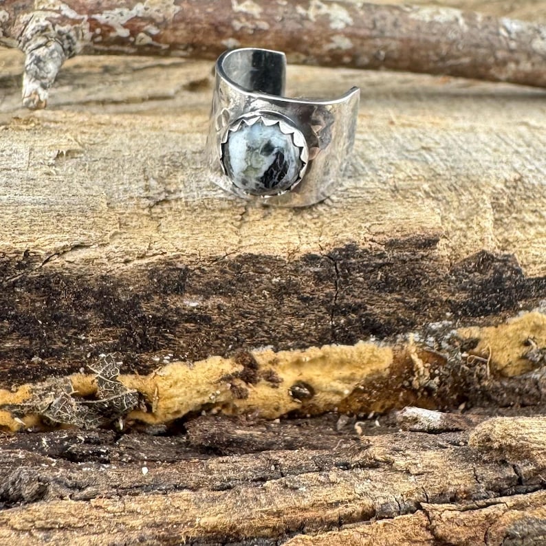 May include: A silver ring with a hammered texture and a round, black and white stone. The ring is set against a natural wood background, showcasing the ring's design and craftsmanship. The ring is a statement piece.