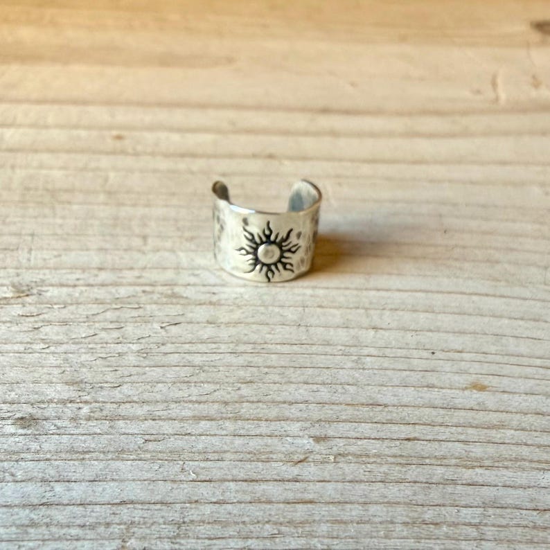 May include: A silver-colored metal ear cuff featuring a hammered texture and a stamped sun design with radiating rays. The open-band accessory has a polished finish and a curved shape, designed to sit securely on the outer edge of the ear cartilage.