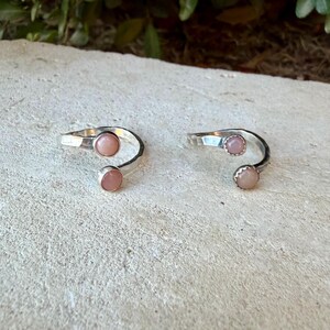 May include: Two open-style silver rings, each featuring two round, smooth, pale pink cabochon stones set at the ends of the band. The silver bands have a polished finish and a bypass design, allowing for an adjustable fit on the finger.
