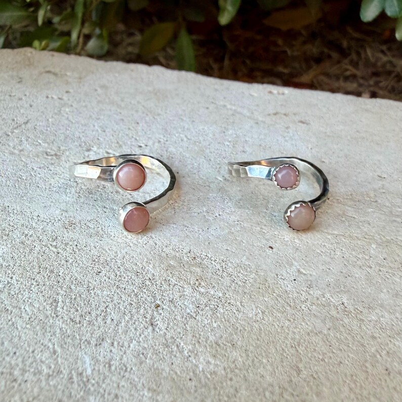 May include: Two open-style silver rings, each featuring two round, smooth, pale pink cabochon stones set at the ends of the band. The silver bands have a polished finish and a bypass design, allowing for an adjustable fit on the finger.