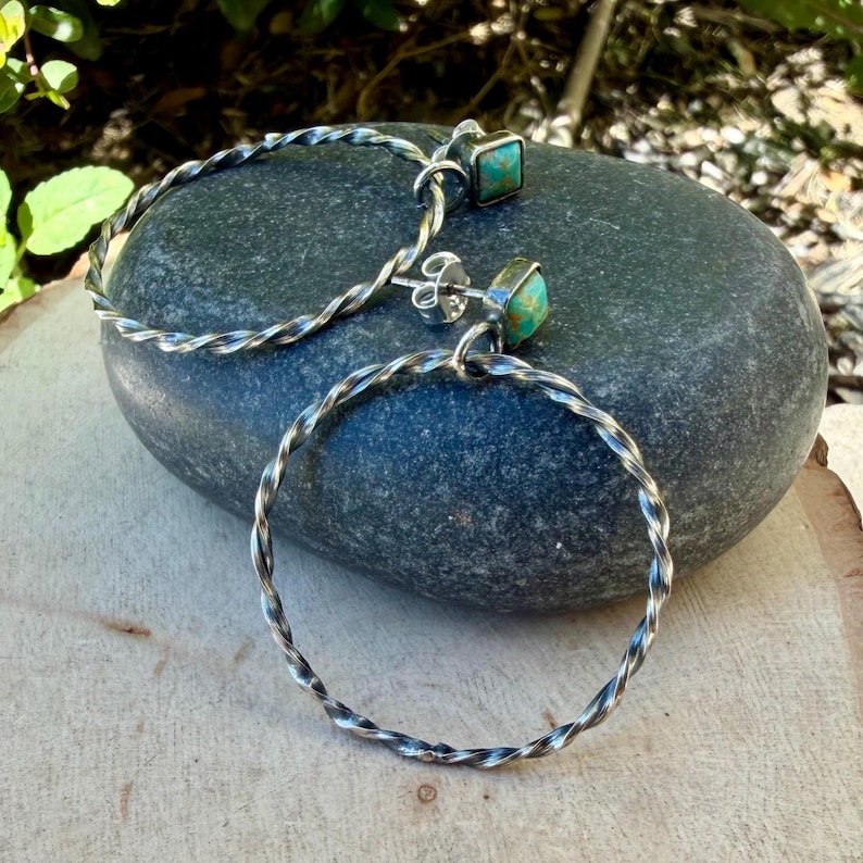May include: A pair of silver hoop earrings with a twisted rope design. Each earring features a small, square turquoise stone. The earrings are displayed on a dark gray stone, showcasing their intricate details and craftsmanship.