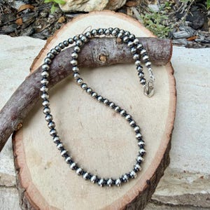 Pu&ograve; includere: Una collana di perline d'argento con chiusura a moschettone. La collana &egrave; composta da numerose piccole perline rotonde d'argento. La collana &egrave; esposta su una superficie di legno con un'estetica naturale e rustica.