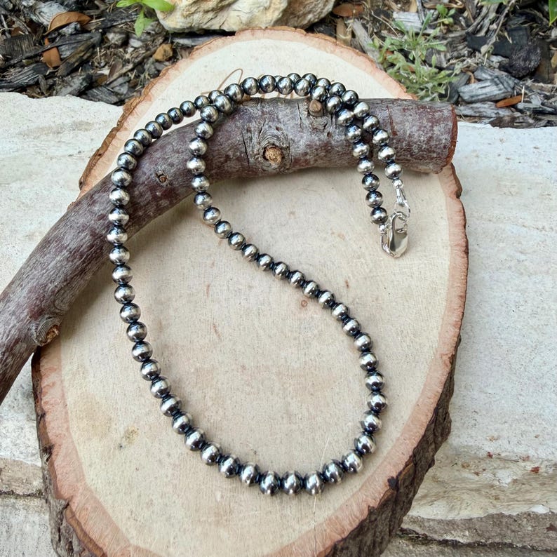 Pu&ograve; includere: Una collana di perline d'argento con chiusura a moschettone. La collana &egrave; composta da numerose piccole perline rotonde d'argento. La collana &egrave; esposta su una superficie di legno con un'estetica naturale e rustica.