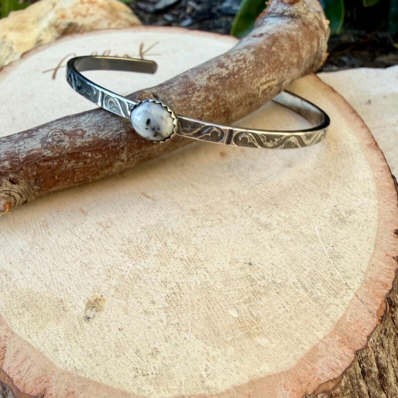 May include: A silver cuff bracelet with an oval, white stone at the centre. The bracelet has an etched design along the band. The stone has a speckled appearance. The bracelet is displayed on a wooden surface.