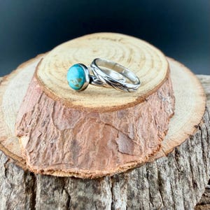 May include: A silver ring set featuring a turquoise stone ring and a matching band with a woven design. The rings are displayed on a rustic wooden surface, showcasing the jewelry's details and craftsmanship. The turquoise stone has a light blue color.