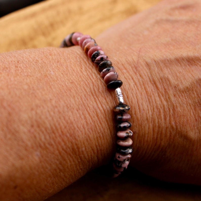 May include: A beaded bracelet featuring pink and black stones. The bracelet is strung on a silver-colored wire. The stones are round and flat, with a mix of pink and black colors. The bracelet is worn on a wrist.
