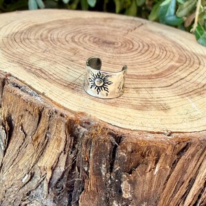 May include: A silver-toned metal ring featuring an open band design. The surface of the ring is engraved with a detailed sun motif, characterized by a central circular core surrounded by radiating, pointed rays that extend across the band.