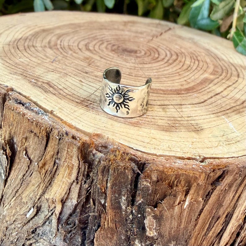 May include: A silver-toned metal ring featuring an open band design. The surface of the ring is engraved with a detailed sun motif, characterized by a central circular core surrounded by radiating, pointed rays that extend across the band.