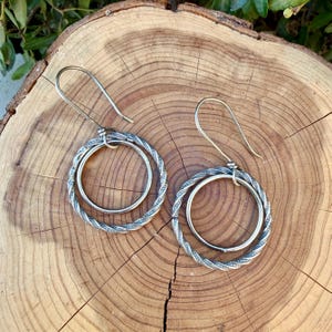 May include: A pair of silver-toned dangle earrings featuring a double-hoop design. Each earring consists of a smooth inner circle suspended within a larger, textured, twisted-wire outer hoop, attached to a simple curved ear wire hook.
