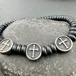 May include: Black beaded bracelet featuring three silver-toned cross pendants. The bracelet is made of matte black beads and has a simple, elegant design. The crosses are detailed with a classic Christian cross design.
