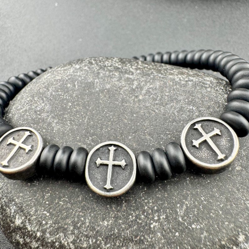 May include: Black beaded bracelet featuring three silver-toned cross pendants. The bracelet is made of matte black beads and has a simple, elegant design. The crosses are detailed with a classic Christian cross design.
