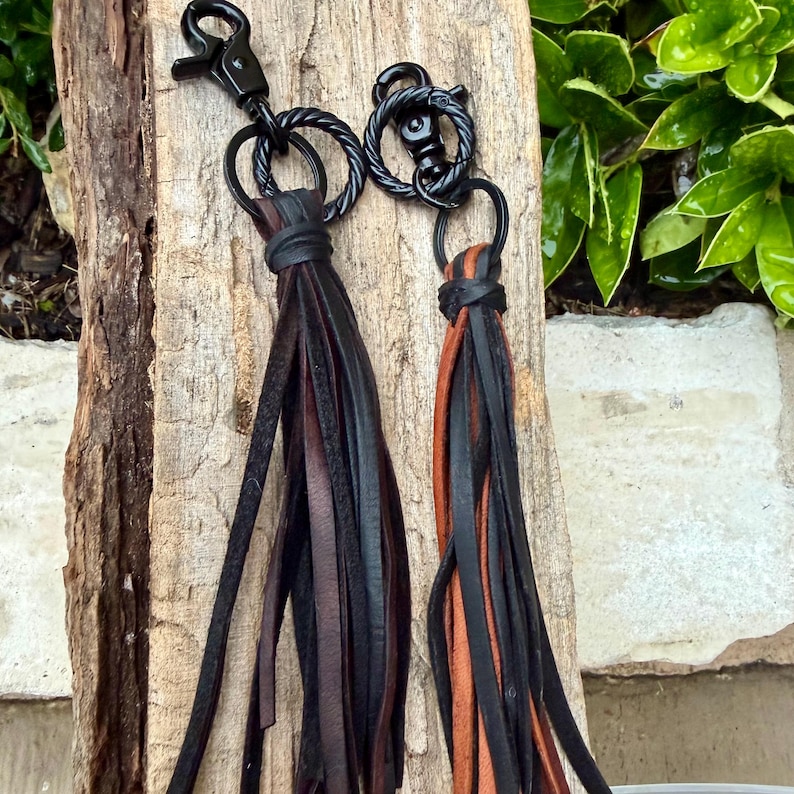 May include: Two leather tassel keychains with black hardware. One keychain has black and brown tassels, and the other has black and orange tassels. The keychains are displayed on a piece of wood.