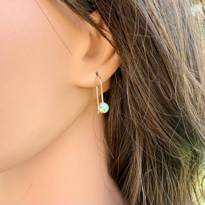 May include: A gold-toned earring featuring a thin, U-shaped wire hook that suspends a small, round charm. The charm displays a mottled, multi-colored surface with flecks of blue, green, and gold, creating a textured appearance on the circular pendant.