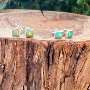 May include: A pair of square stud earrings featuring natural turquoise stones set in silver. Each earring displays a unique blue-green color with brown matrix veining, secured with a silver post backing for a clean and simple look.