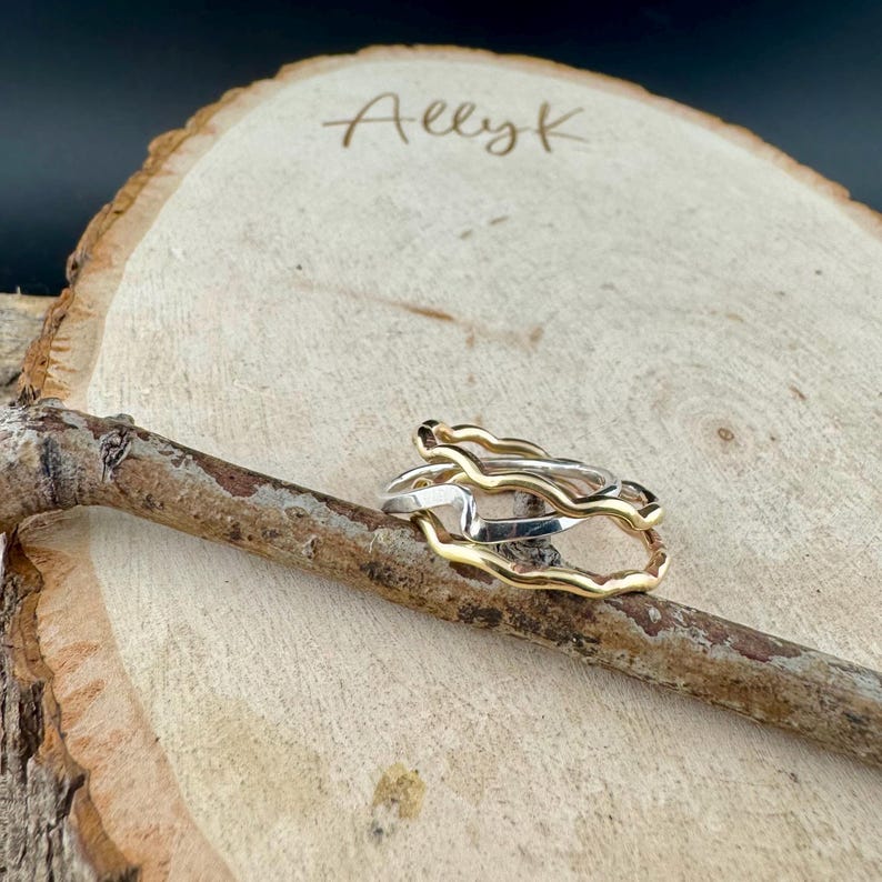 Puede incluir: Tres anillos, dos dorados y uno plateado, apilados sobre una superficie de madera. Los anillos tienen un dise&ntilde;o org&aacute;nico y ondulado. La palabra "Allyk" est&aacute; escrita en cursiva sobre la madera.