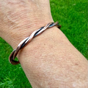 May include: A copper-toned bracelet with a twisted design. The bracelet is made of two strands of metal that are intertwined. The bracelet is worn on a wrist, with a green background.
