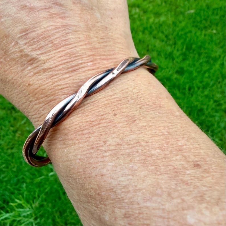 May include: A copper-toned bracelet with a twisted design. The bracelet is made of two strands of metal that are intertwined. The bracelet is worn on a wrist, with a green background.