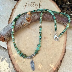 May include: A turquoise beaded necklace with a silver feather pendant. The necklace features small, round beads in shades of green and brown. The feather pendant hangs from a silver metal accent. The necklace is displayed on a wooden surface.