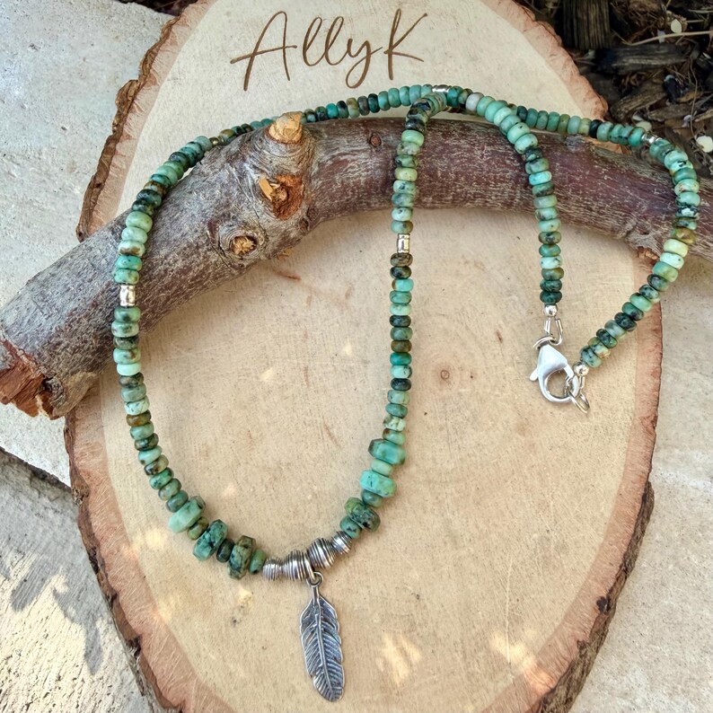 May include: A turquoise beaded necklace with a silver feather pendant. The necklace features small, round beads in shades of green and brown. The feather pendant hangs from a silver metal accent. The necklace is displayed on a wooden surface.