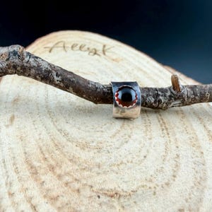 Puede incluir: Un anillo de plata con una piedra preciosa de color rojo oscuro engastada en una banda de metal martillado. El anillo se muestra sobre una rama de madera sobre un fondo de madera clara. La piedra es redonda y brillante.