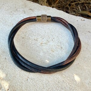 May include: A circular bracelet composed of two intertwined leather cords, one in black and one in dark brown. The cords are secured together by a cylindrical, metallic brass-toned clasp with a smooth finish, creating a simple and functional design.