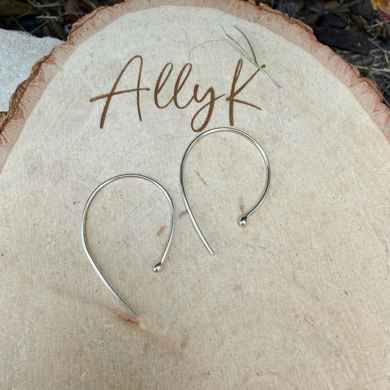 Puede incluir: Un par de pendientes de aro plateados con un dise&ntilde;o curvo y abierto con peque&ntilde;os extremos esf&eacute;ricos. Los pendientes est&aacute;n colocados sobre una superficie de madera clara que presenta el texto grabado Ally K en letra cursiva.