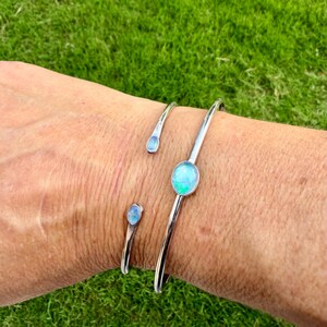 K&ouml;nnte beinhalten: Drei silberfarbene Armb&auml;nder mit blau-gr&uuml;nen, opalartigen Steinen. Die Armb&auml;nder werden an einem Handgelenk vor dem Hintergrund von gr&uuml;nem Gras getragen. Die Armb&auml;nder haben ein minimalistisches Design, wobei die Steine im Mittelpunkt stehen.