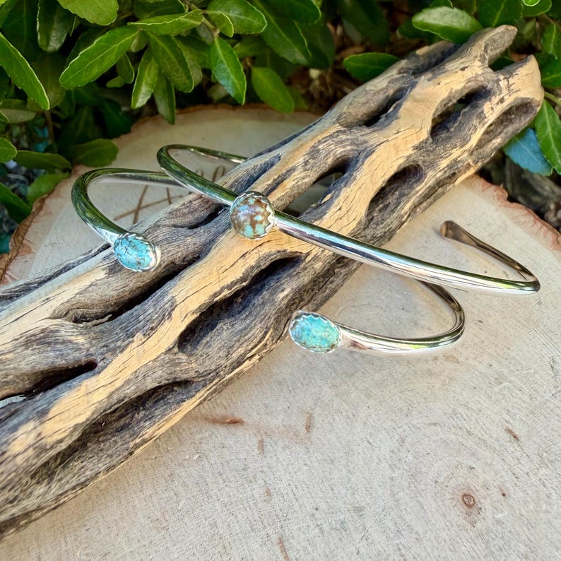 May include: Two silver-toned cuff bracelets with turquoise-colored stones. The bracelets are displayed on a piece of weathered wood, with green foliage in the background. The stones are oval-shaped and set in a silver bezel.