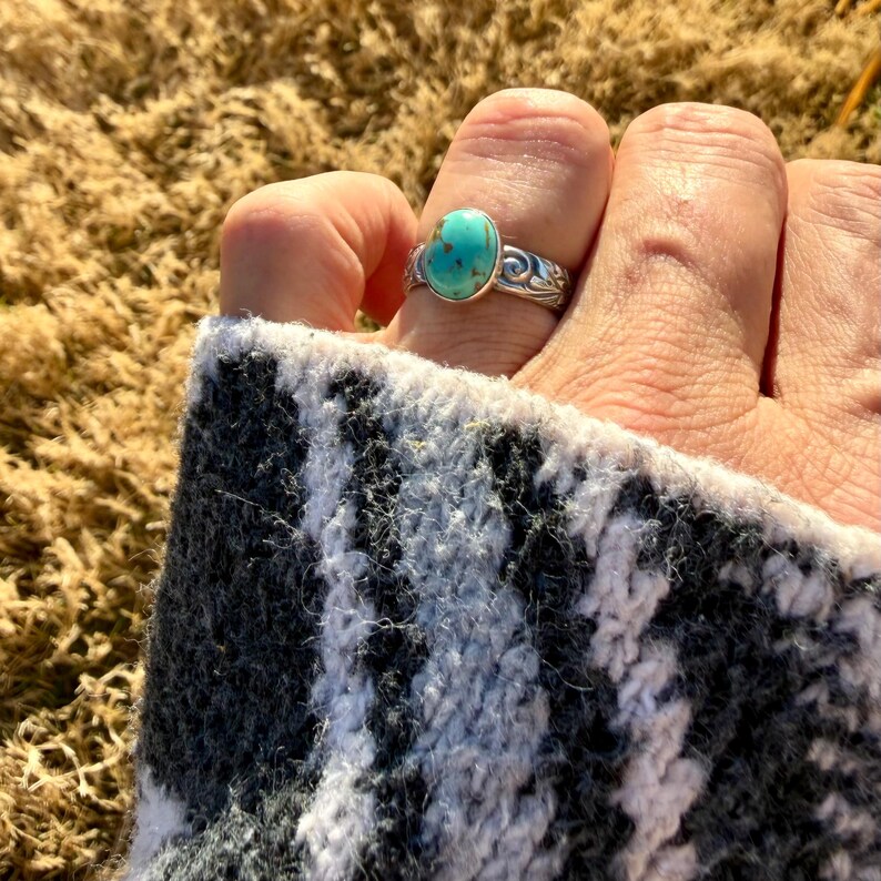 May include: A silver ring with an oval turquoise stone. The ring has a decorative, swirling pattern around the stone. The ring is on a finger, and the person is wearing a black and white patterned sweater.