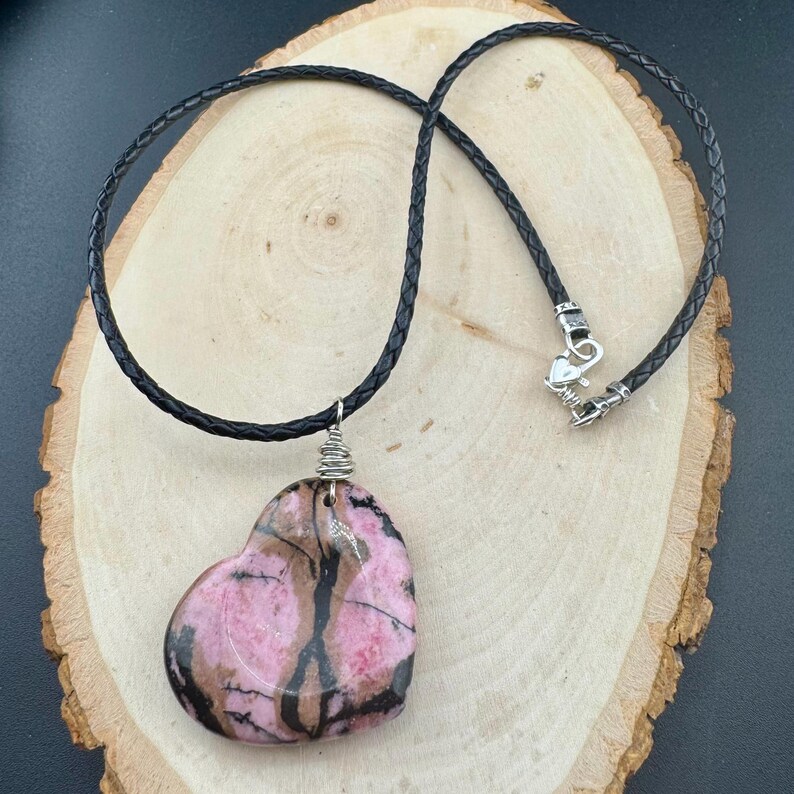 May include: A heart-shaped pendant necklace featuring a pink and brown stone. The stone is wire-wrapped and suspended from a black braided cord with a silver-toned clasp. The necklace rests on a wooden surface.