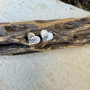 May include: A pair of silver heart-shaped stud earrings featuring an intricate, embossed scrollwork pattern across the surface. The metal has a polished finish with darkened recessed areas that highlight the detailed swirling design.