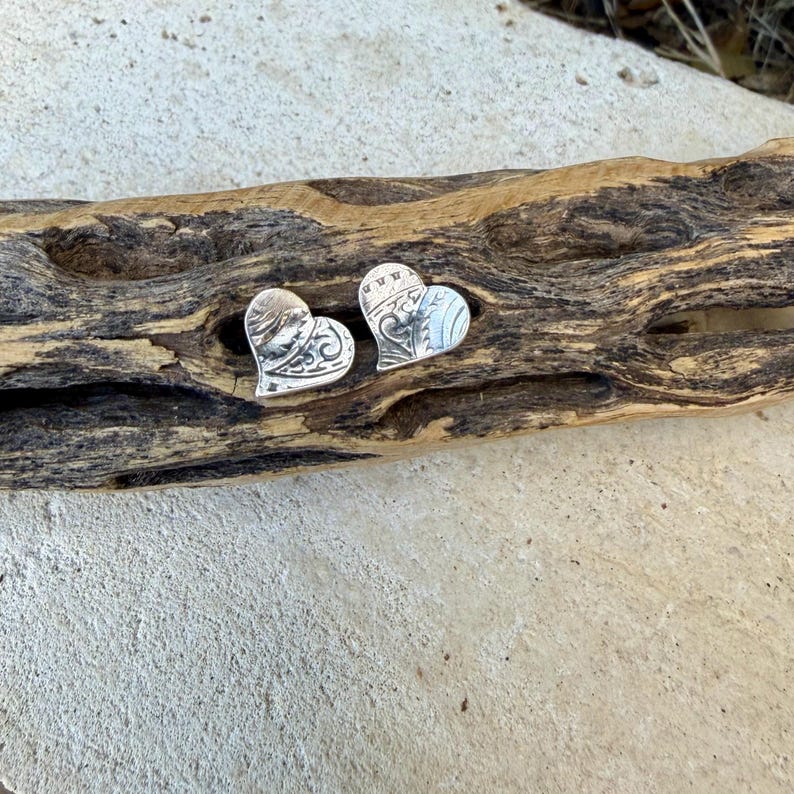 May include: A pair of silver heart-shaped stud earrings featuring an intricate, embossed scrollwork pattern across the surface. The metal has a polished finish with darkened recessed areas that highlight the detailed swirling design.