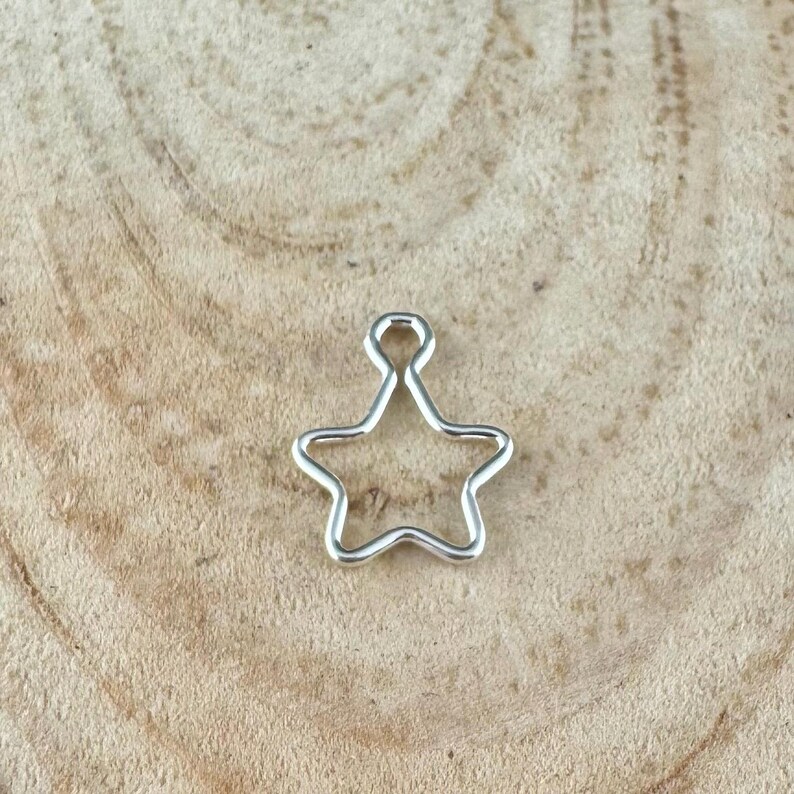 May include: A silver-colored star-shaped charm with a loop at the top for hanging. The charm is made of a shiny metal and has a simple, open design. The star is displayed on a light brown wooden surface.