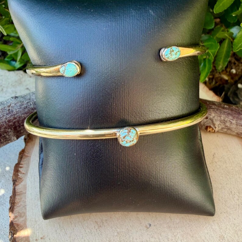 May include: Two gold-colored metal bracelets with turquoise-colored stones. One bracelet is a cuff style, and the other is a simple band. The bracelets are displayed on a black surface.