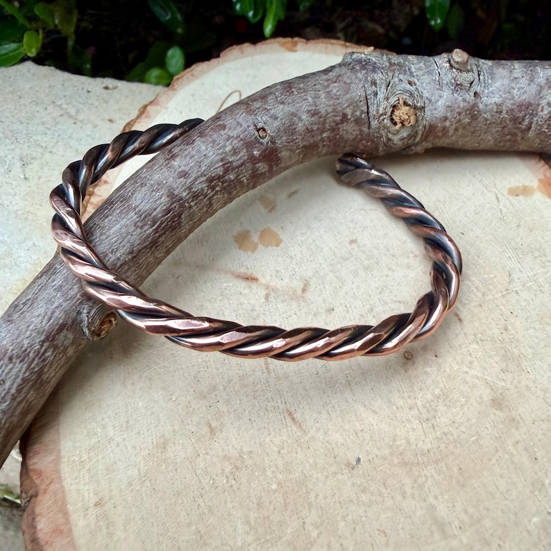 May include: A copper-colored twisted metal bracelet. The bracelet is a solid band with a textured, braided appearance. The metal has a warm, reddish-brown hue with darker accents, resting on a wooden surface.