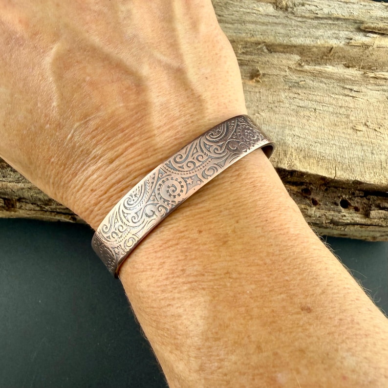 Puede incluir: Un brazalete de color cobre con un intrincado patr&oacute;n repetitivo de remolinos y dise&ntilde;os florales. El brazalete de banda ancha se lleva en la mu&ntilde;eca, mostrando la detallada obra de arte en relieve. El metal tiene un tono c&aacute;lido, marr&oacute;n rojizo.