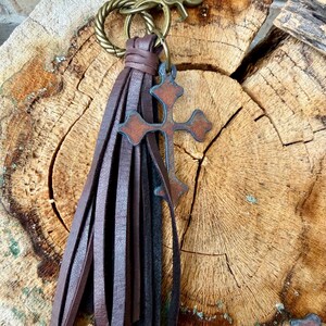 Op de afbeelding: Een donkerbruine leren kwast-sleutelhanger met een decoratieve metalen kruisbedel met een roestige afwerking. De kwast is bevestigd aan een gedraaide messingkleurige metalen ring en een karabijnhaak, een functioneel accessoire.