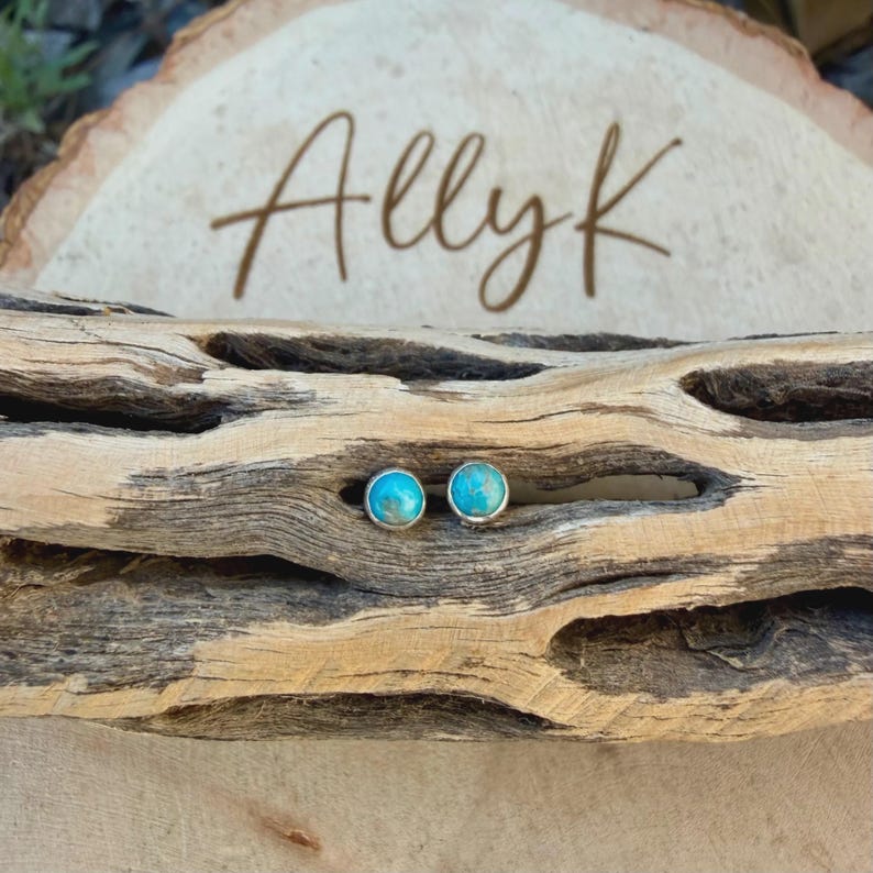 May include: A pair of turquoise stud earrings with silver settings. The earrings are displayed on a piece of driftwood, with the name "Allyk" written in cursive on a wooden background.