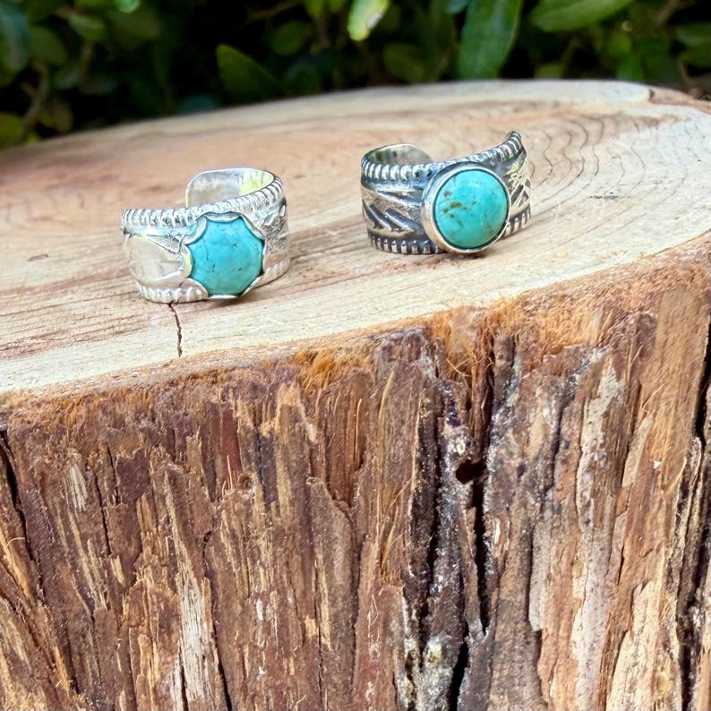 May include: Two silver rings featuring round turquoise stones set in metal bands. The left ring has a scalloped edge around the stone, while the right ring features a smooth bezel setting. Both bands display etched patterns along the sides.