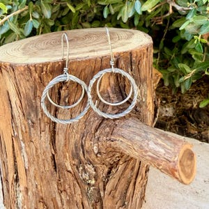 May include: A pair of silver-toned dangle earrings featuring a double-hoop design. Each earring consists of a larger, twisted-texture outer hoop and a smaller, smooth inner hoop, suspended from a simple silver-toned hook wire finding.