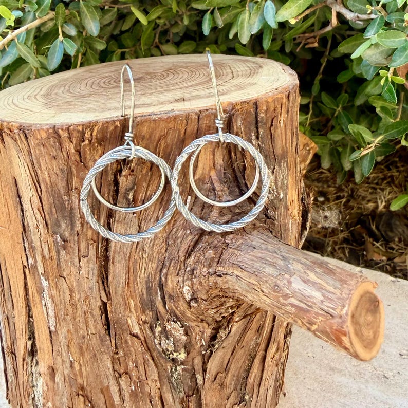 May include: A pair of silver-toned dangle earrings featuring a double-hoop design. Each earring consists of a larger, twisted-texture outer hoop and a smaller, smooth inner hoop, suspended from a simple silver-toned hook wire finding.