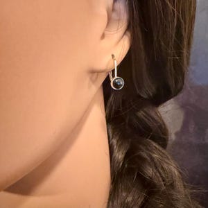 May include: A silver-toned leverback earring with a small, round, black stone. The earring is shown on a person's ear, with dark hair visible in the background. The earring is a simple, elegant design.