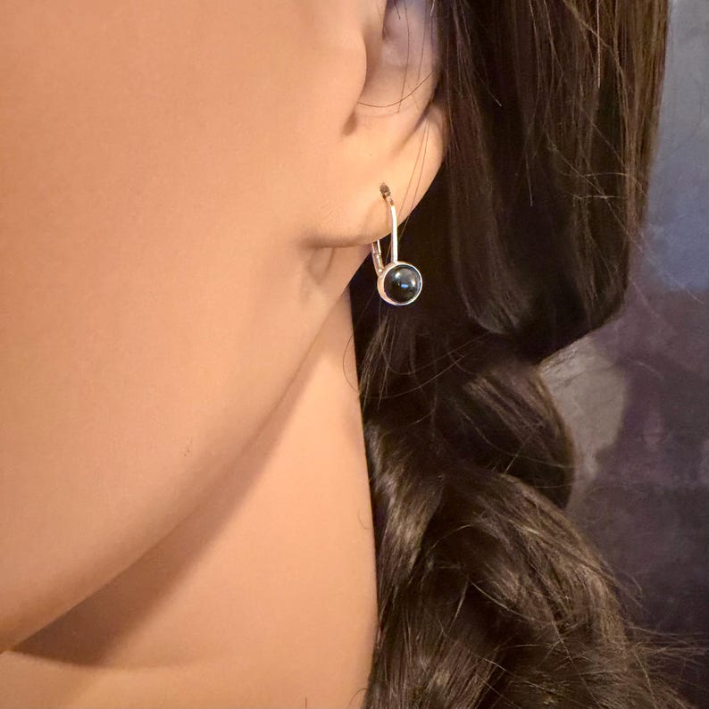 May include: A silver-toned leverback earring with a small, round, black stone. The earring is shown on a person's ear, with dark hair visible in the background. The earring is a simple, elegant design.