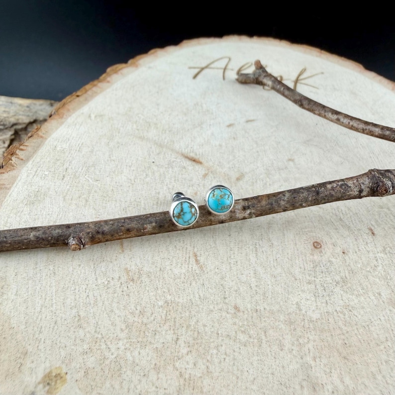May include: A pair of round stud earrings with turquoise stone centers. The earrings are set in silver-colored metal. The earrings are displayed on a wooden surface with a small branch.
