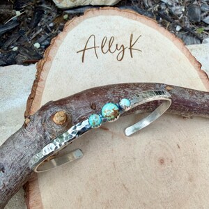 May include: A silver cuff bracelet with a hammered texture and three turquoise stones. The bracelet is displayed on a wooden surface with the name "Allyk" written in cursive.