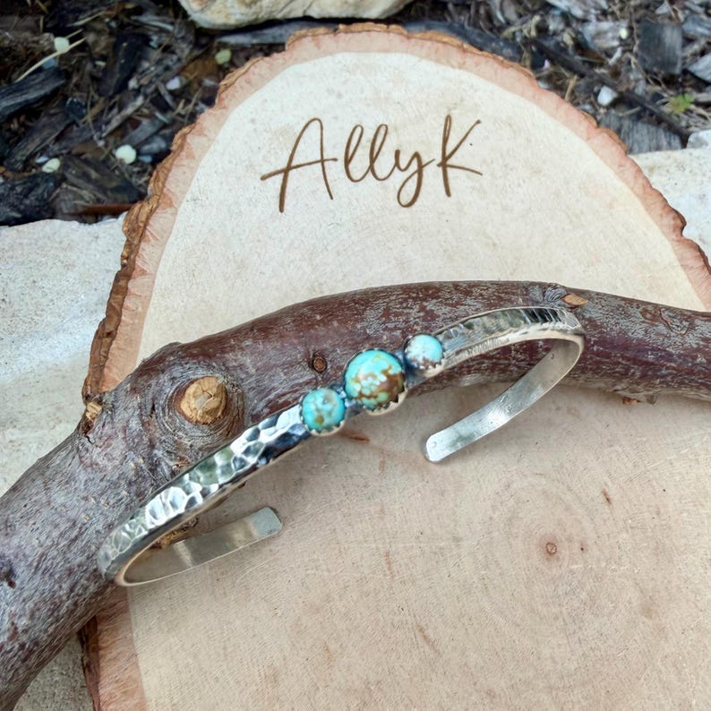 May include: A silver cuff bracelet with a hammered texture and three turquoise stones. The bracelet is displayed on a wooden surface with the name "Allyk" written in cursive.