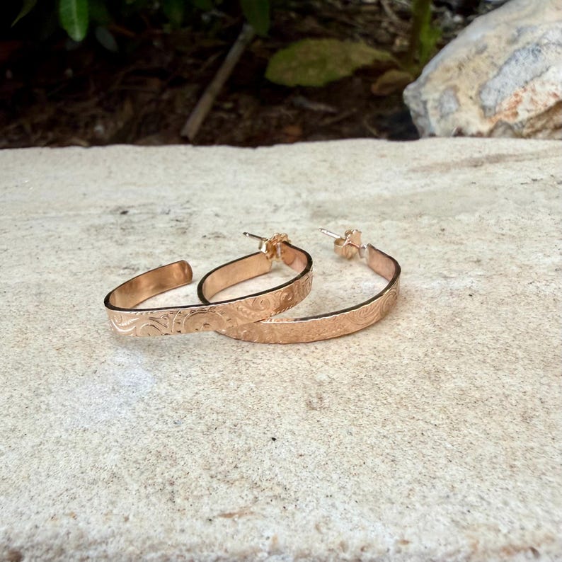May include: A pair of rose gold hoop earrings with an engraved floral pattern. The earrings are displayed on a light-colored, textured surface. The earrings have a classic hoop design with a post closure.
