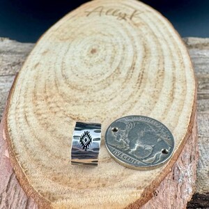 May include: A silver ring with a geometric black design and a buffalo nickel coin on a wooden surface. The ring has a textured surface and a rectangular shape. The coin is silver and has the words "FIVE CENTS" on it.