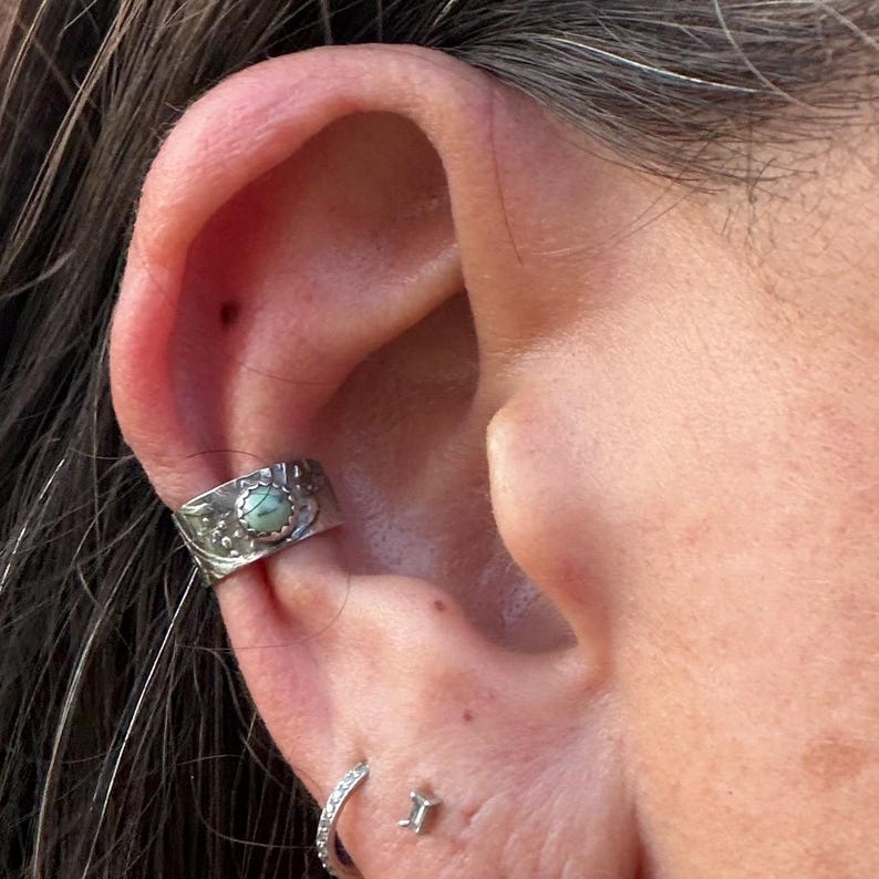 May include: A silver ear cuff with a turquoise stone set in the center. The cuff is decorated with a floral design and is worn on the upper ear. A small hoop earring and a stud earring are also visible.